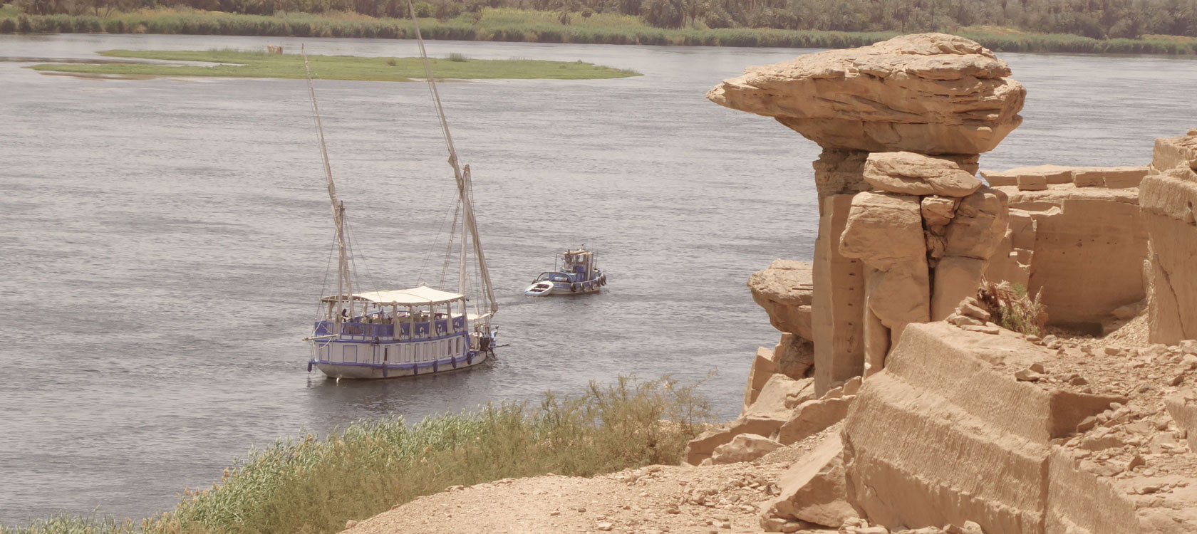 dahabiya-nile-sailing-rock-view View of the Dahabiya Eugenie sailing along the Nile, seen from the cliffs with ancient rock formations in the foreground.